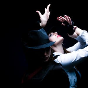 Two individuals, striking dramatic poses while wearing the Porro Neque Quisq black hats and dark clothing, are set against a black background, with one gazing upward and the other looking downward.