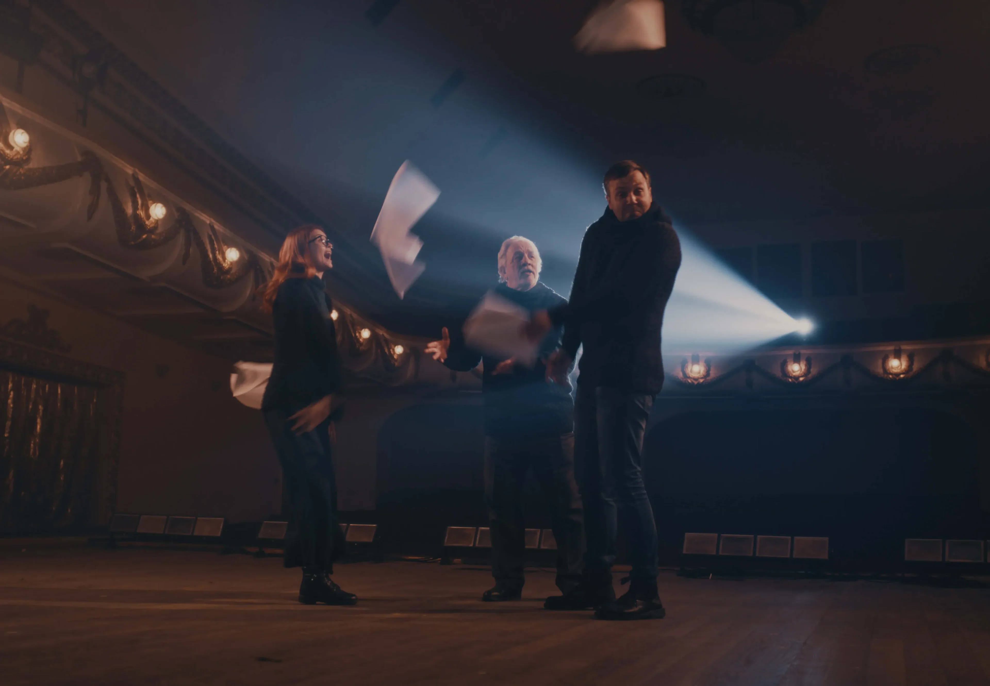 Three individuals on a dimly lit stage toss papers into the air, highlighted by a spotlight shining behind them, showcasing the Neque Porro Quisq.