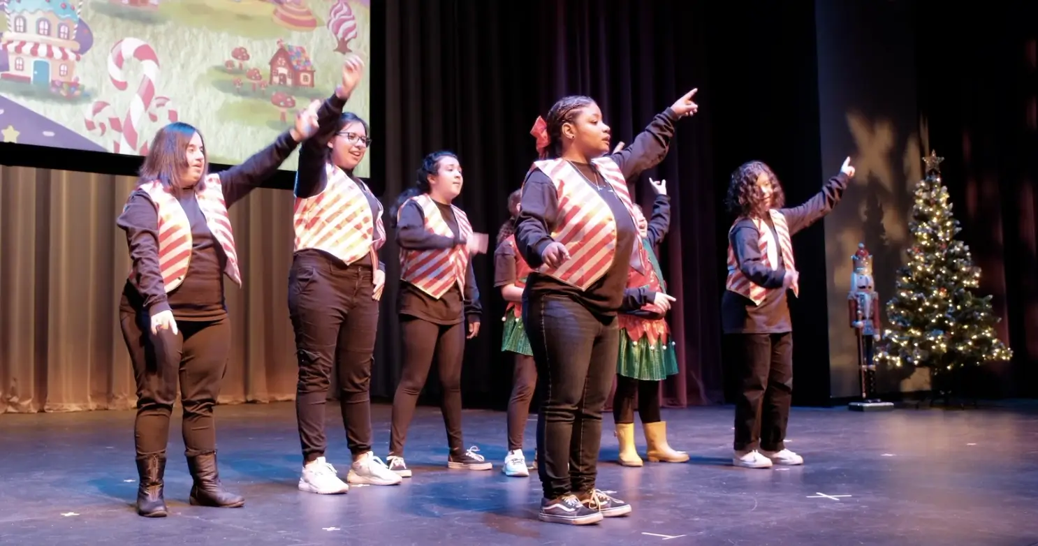 Performers on stage pointing, holiday backdrop.